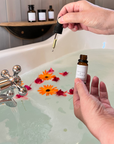 Person holding a dropper over a bathtub filled with water and flowers, with a bottle in the foreground.