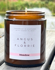Candle in a brown jar with 'Angus & Florrie' label on a wooden surface with a blurred natural background