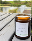 Candle in a brown jar with a label on a wooden surface, outdoors.