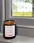 Candle with 'Angus & Florrie' label on a windowsill with a view of trees outside.