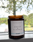 Candle in an amber jar labeled 'Angus & Florrie' on a windowsill with a blurred natural background