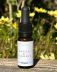 Bottle of Angus & Florrie Tranquil oil with a black dropper on a wooden surface with flowers in the background