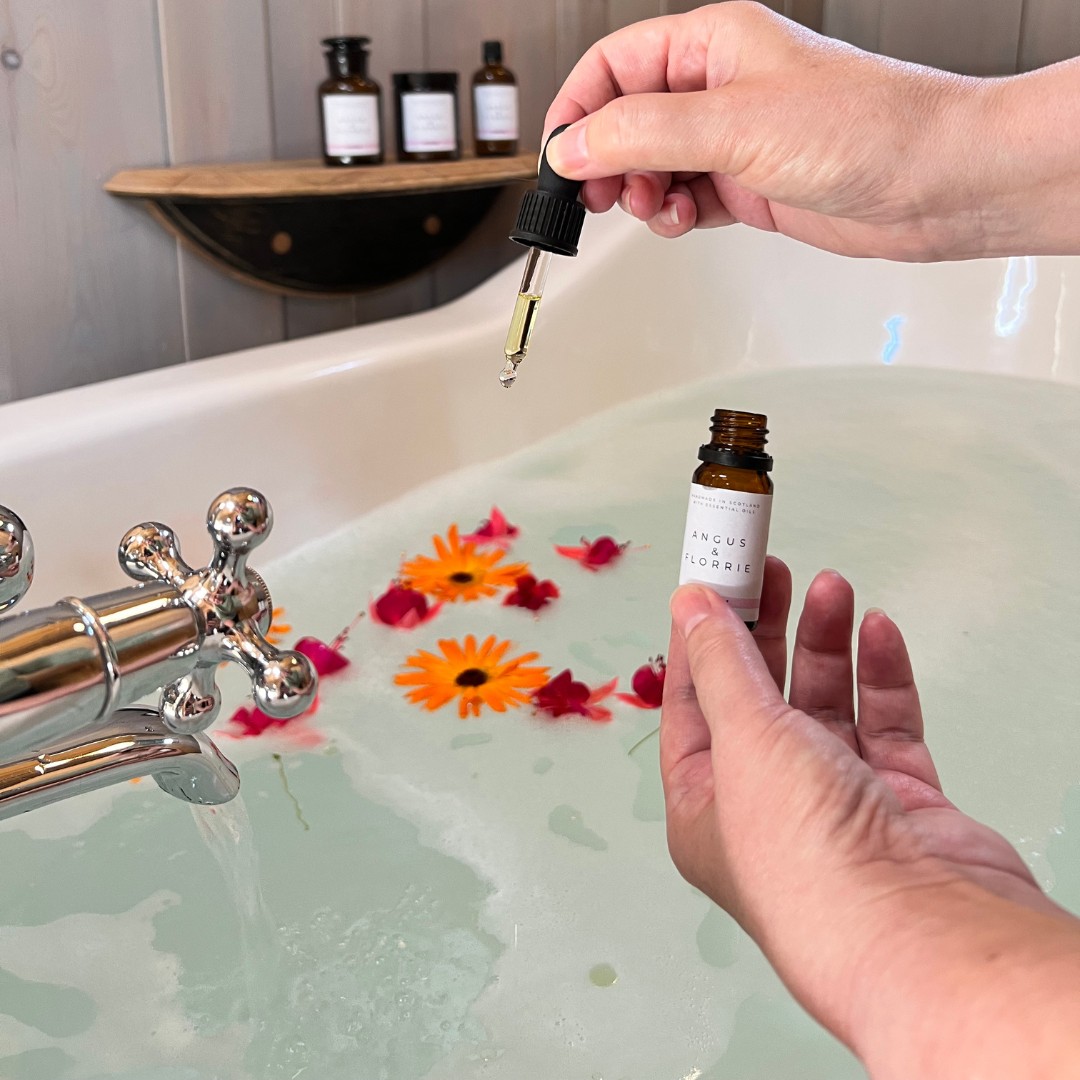 Person holding a dropper over a bathtub filled with water and flowers, with a bottle in the foreground.