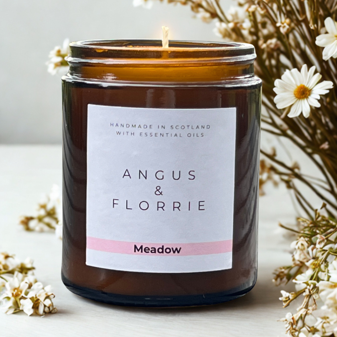 Candle in a brown jar with 'Angus & Florrie' label surrounded by flowers