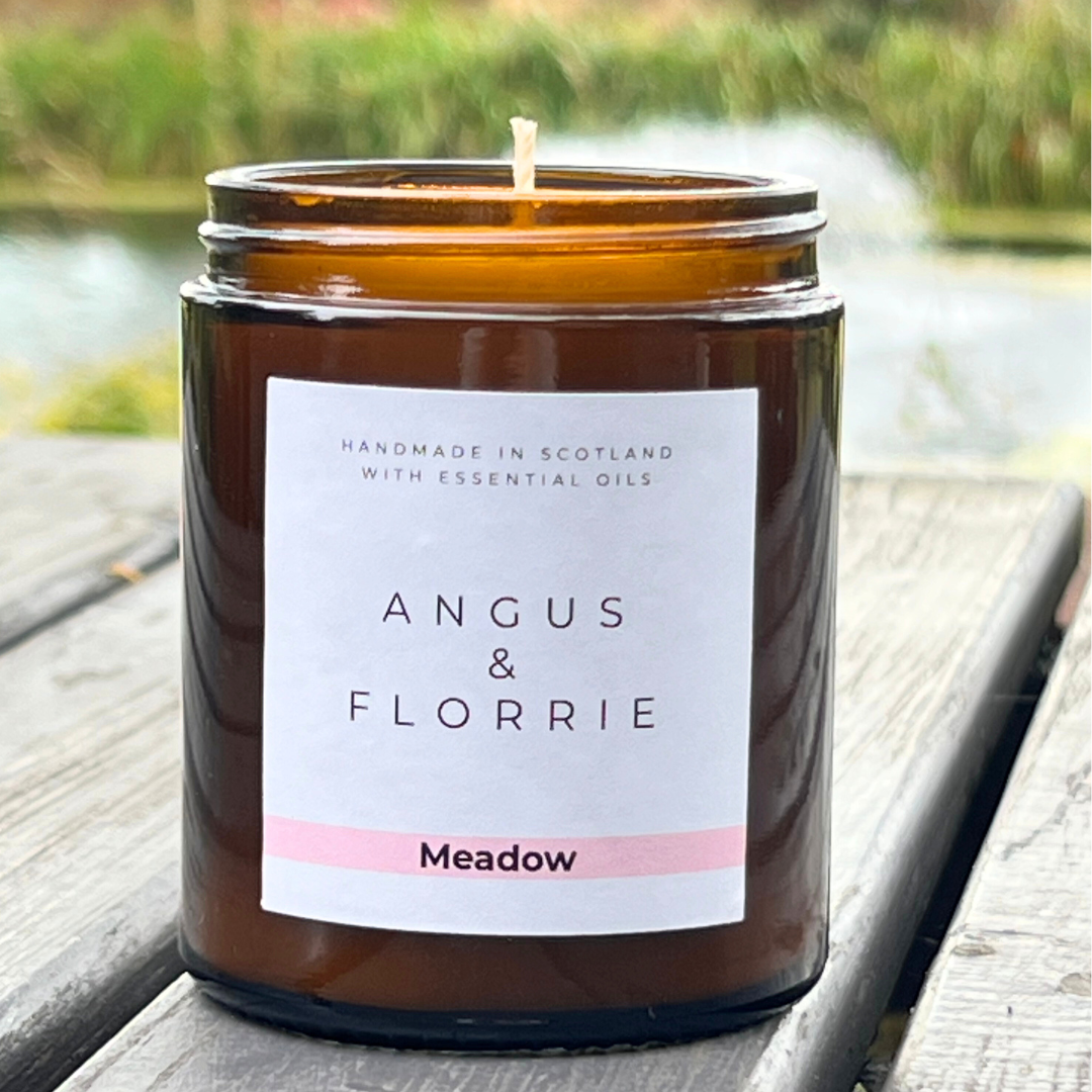 Candle in a brown jar with 'Angus & Florrie' label on a wooden surface with a blurred natural background