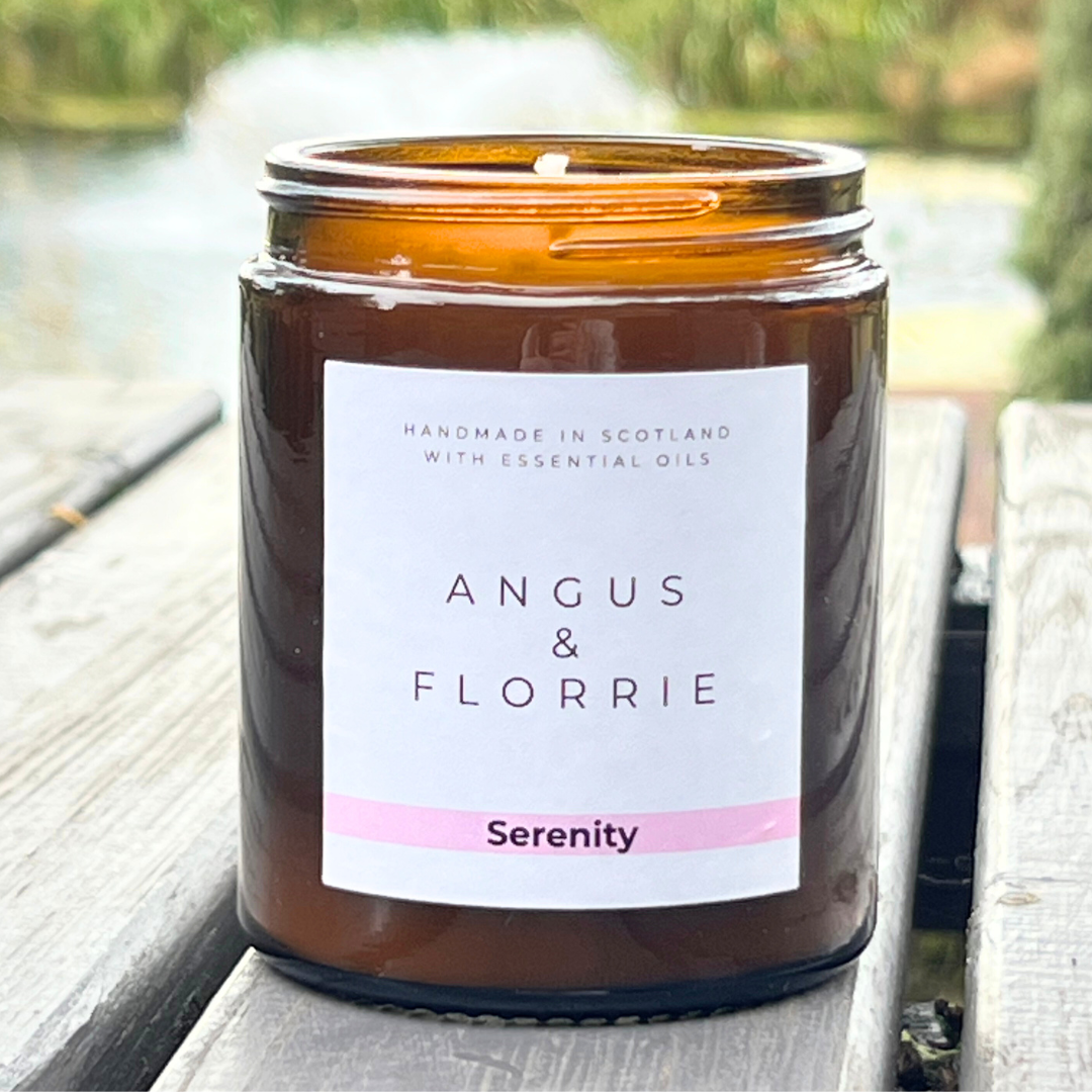 Candle in an amber jar with a label reading 'Angus & Florrie Serenity' on a wooden surface.
