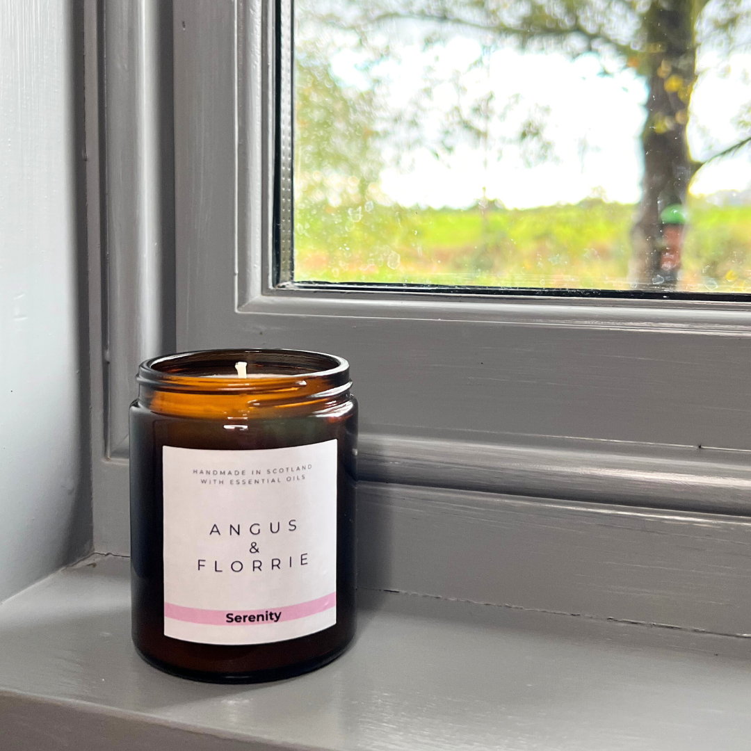 Candle with 'Angus & Florrie' label on a windowsill with a view of trees outside.