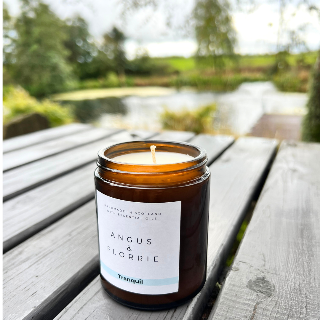 Candle in an amber jar with a label on a wooden surface near a natural landscape