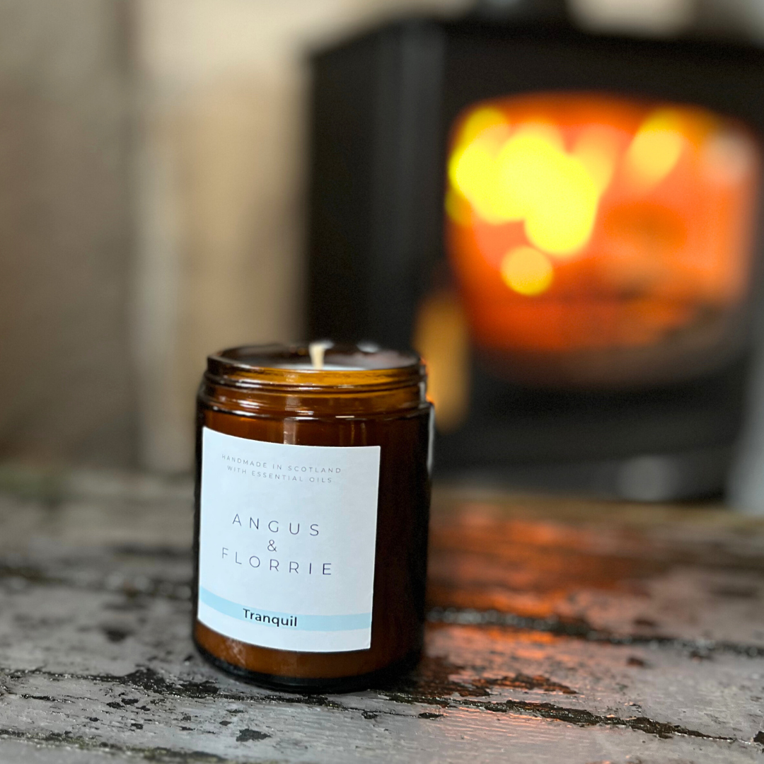 Candle in a brown jar with a label reading 'Angus & Florrie' on a rustic surface with a blurred background.