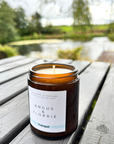 Candle in an amber jar with a label on a wooden surface near a natural landscape