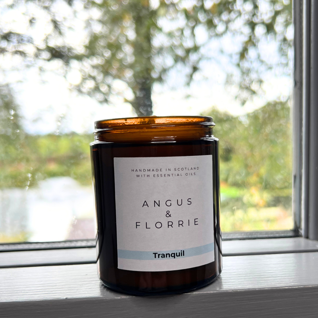 Candle in an amber jar labeled 'Angus & Florrie' on a windowsill with a blurred natural background