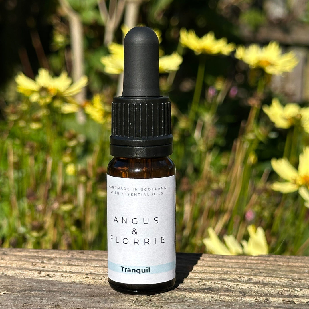Bottle of Angus & Florrie Tranquil oil with a black dropper on a wooden surface with flowers in the background
